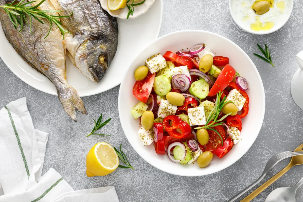 tavolo con un piatto con pesce alla griglia e insalata con pomodorini olive e feta