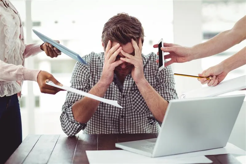 Uomo stressato al pc mentre riceve compiti da fare da altre due persone, tra cui compilare fogli e rispondere al telefono