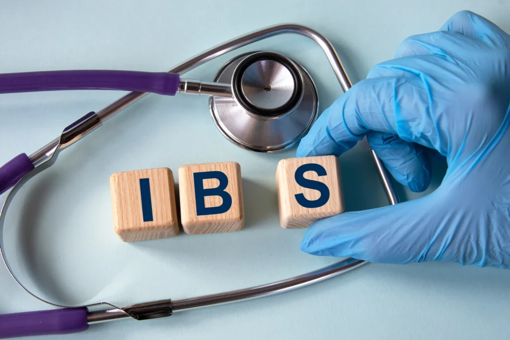 Una mano con un guanto azzurro compone la parola "IBS" usando tre cubi con le rispettive lettere, posizionando la scritta su un tavolo dove già c'è uno stetoscopio.